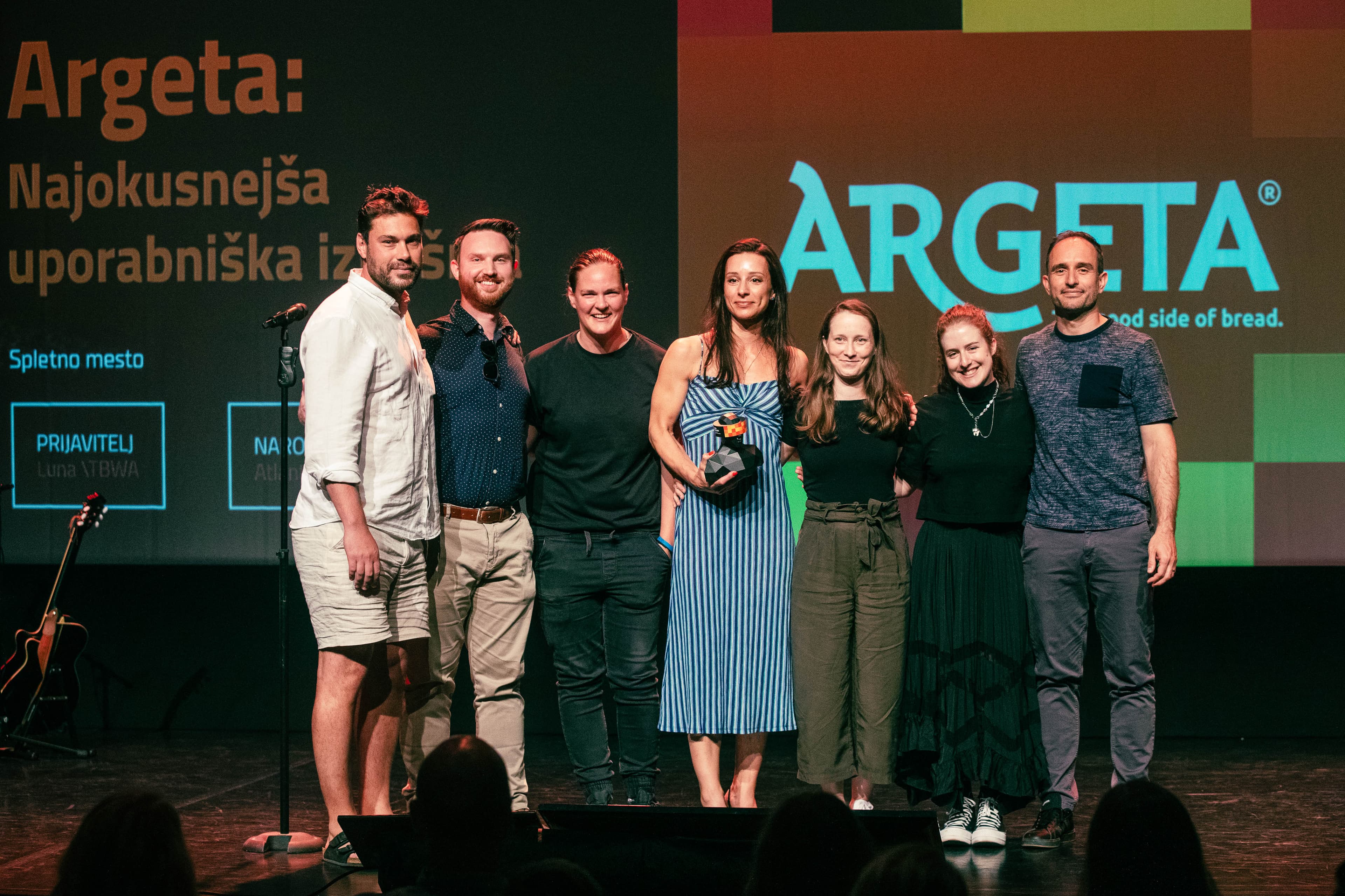 A group of seven people standing together on stage, smiling and holding an award, in front of a large screen displaying the Argeta logo and Slovenian text “Najokusnejša uporabniška izkušnja.” The background features colorful geometric patterns and digital award information.