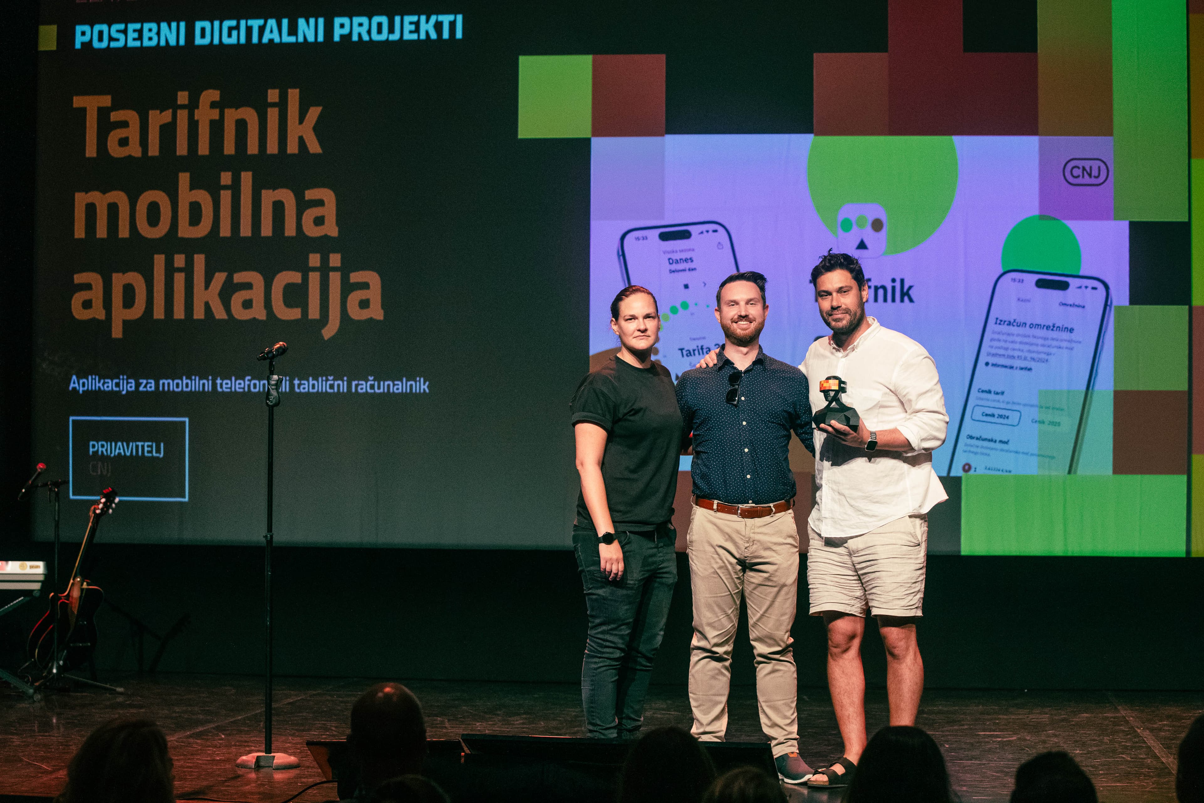 Three people from the CNJ Digital team standing on stage holding an award, with a large screen behind them displaying the title &quot;Tarifnik mobilna aplikacija&quot; and images of the Tarifnik mobile app interface, during a digital project event.