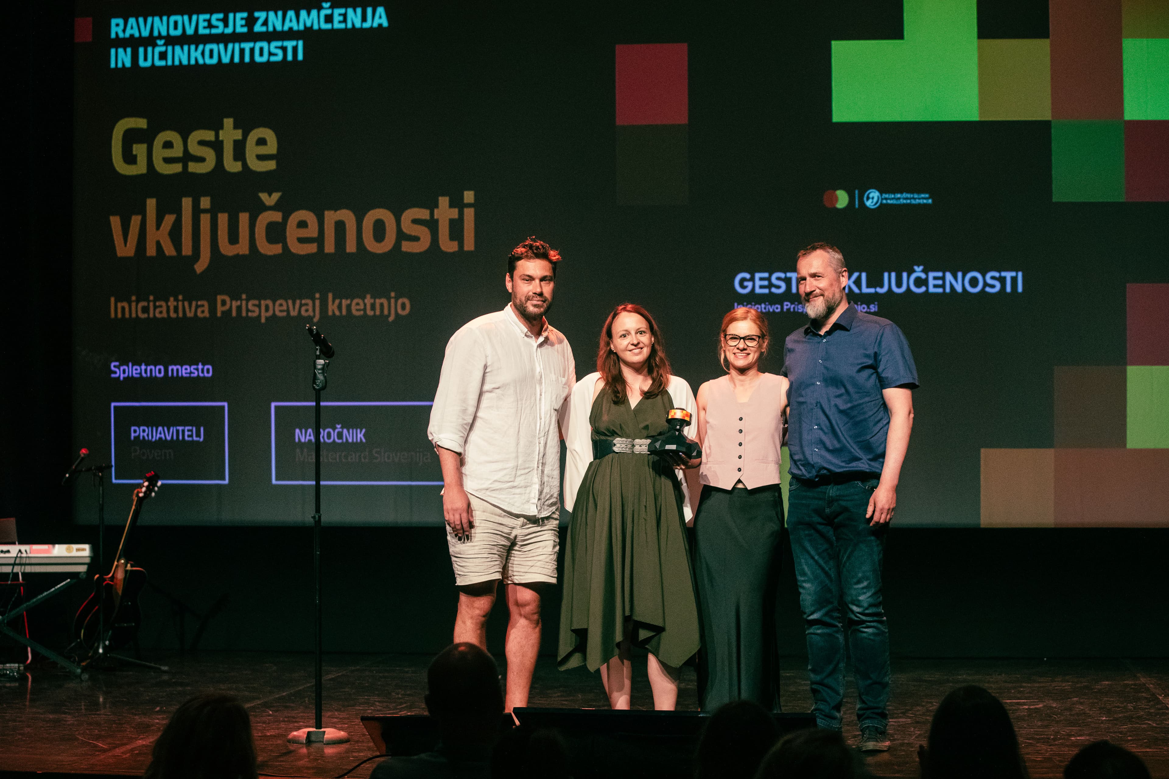 Four people posing together on stage, one holding a trophy, in front of a large screen displaying Slovenian text about inclusion and achievement. The screen reads &quot;Geste vključenosti&quot; and shows options for &quot;Prijatelj&quot; and &quot;Na očeh,&quot; with branding for CNJ Digital visible. Stage lighting highlights the group in a formal event setting.