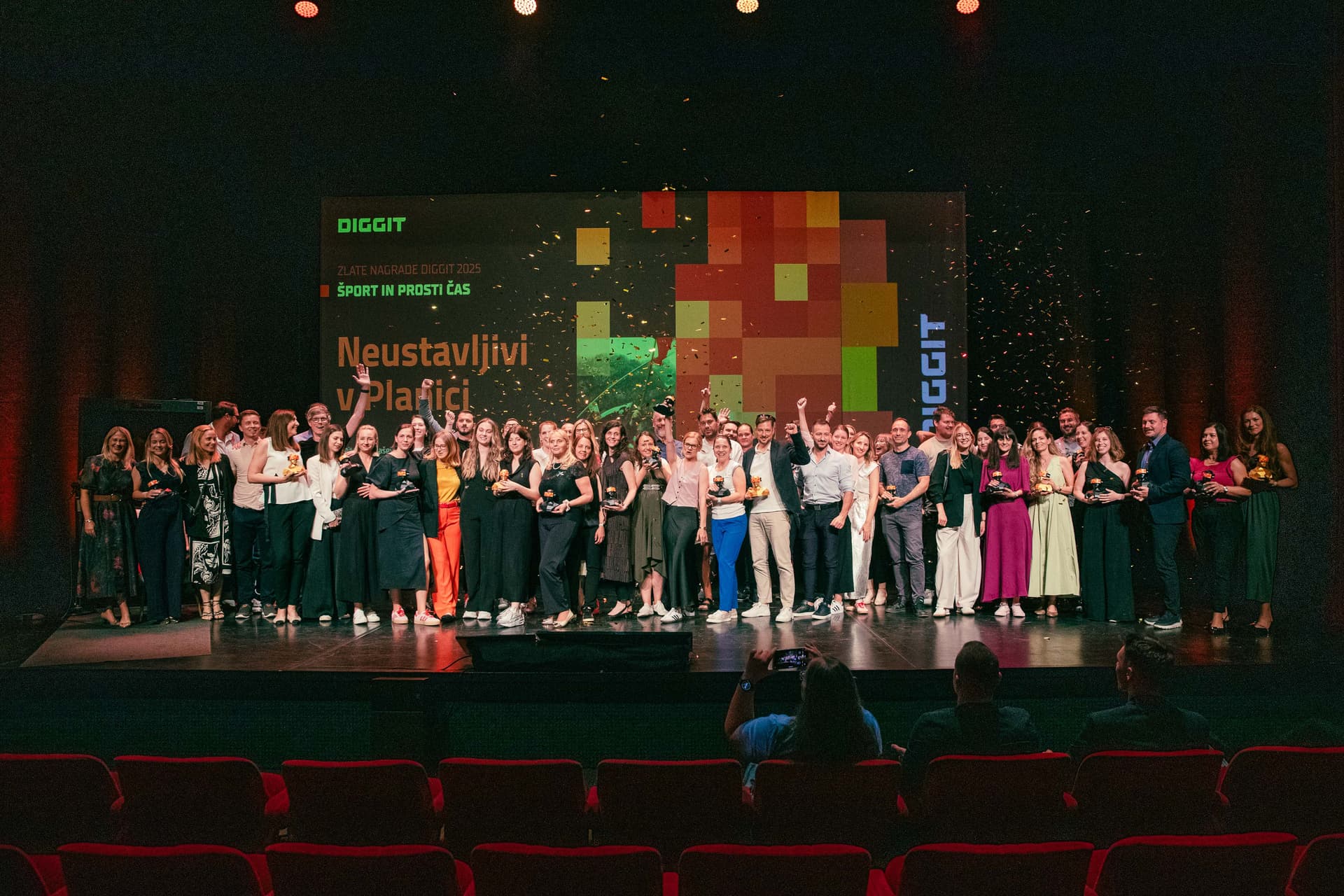 Group of award recipients and presenters posing on stage at the DIGGIT event, backed by a colorful digital-themed screen displaying the text "DIGGIT", "Neustavljivi v Plani", and other Slovenian phrases, with confetti falling and audience members capturing the moment from the front row.