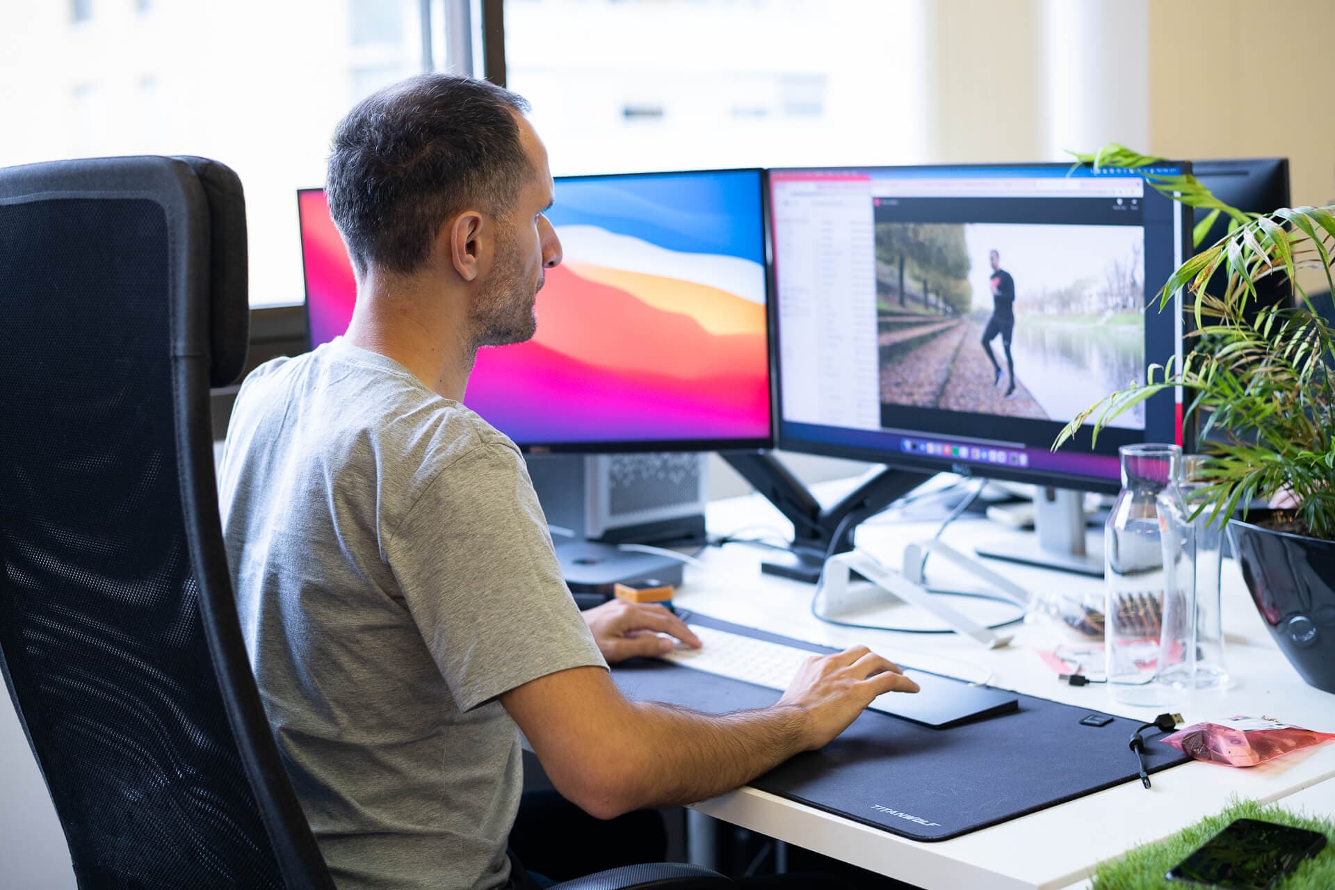 A man is sitting at a desk in an office, focused on his computer monitor. He has two screens in front of him, one displaying a vibrant background and the other showing an image of a person jogging near a body of water. A plant and a glass container are visible on the desk, adding to the workspace aesthetics.