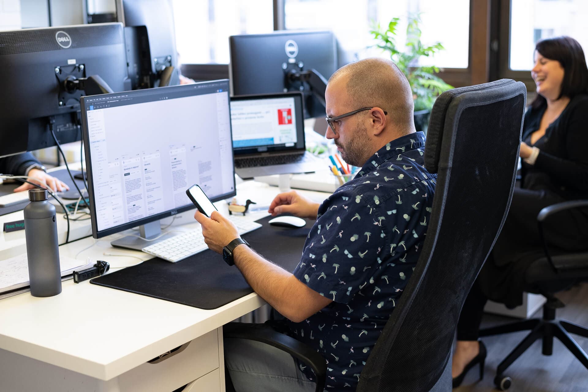 A man with glasses and a patterned shirt sits at a desk, looking at his smartphone. Two computer monitors display various documents, and another person is visible in the background, laughing. A water bottle and other office supplies are scattered on the desk.