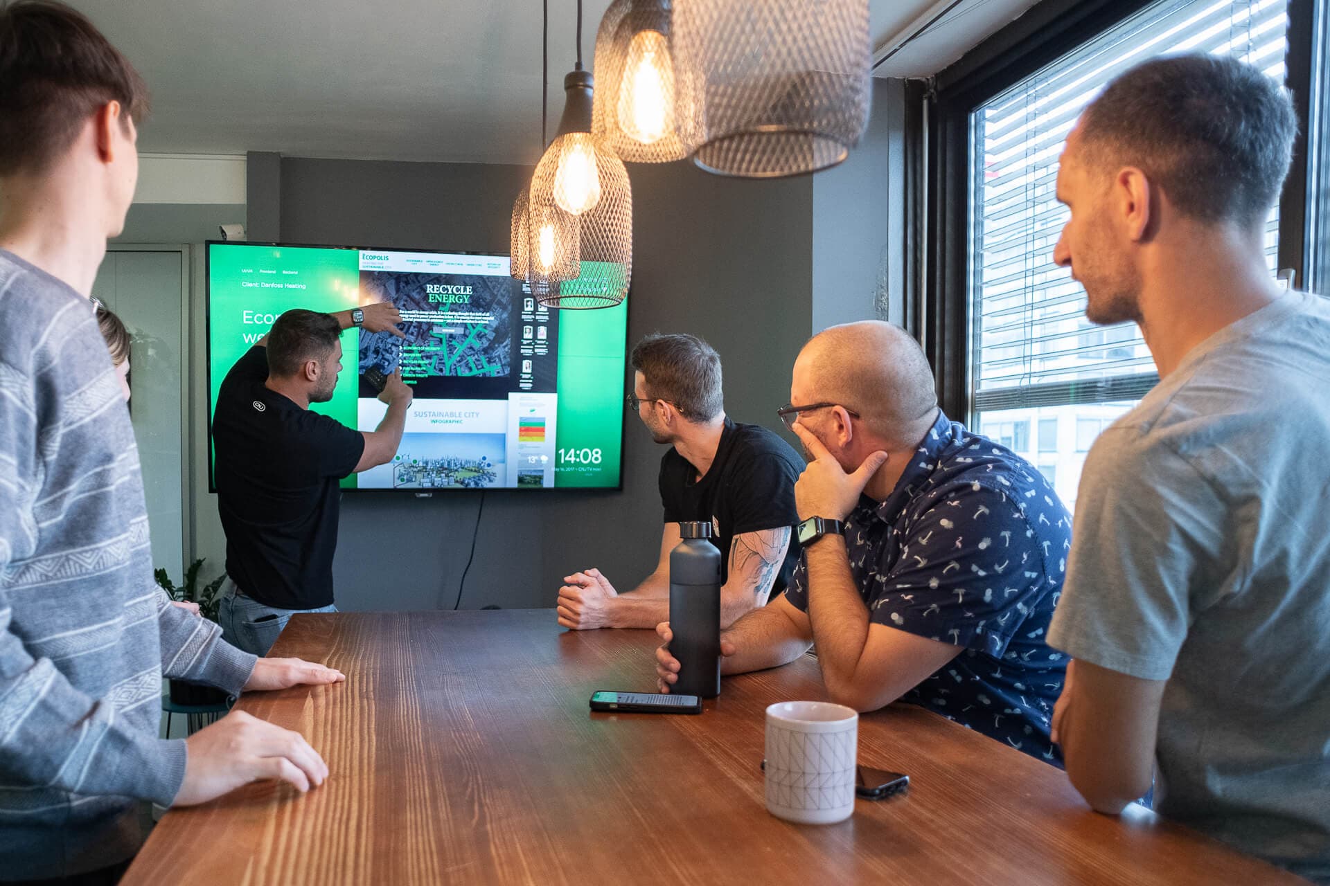 A group of four people are gathered around a conference table, attentively watching a presentation on a large screen. One person is actively pointing at the screen, which displays various graphs and information related to sustainability and energy. The room is well-lit with pendant lights, and there are drinks and electronic devices on the table.