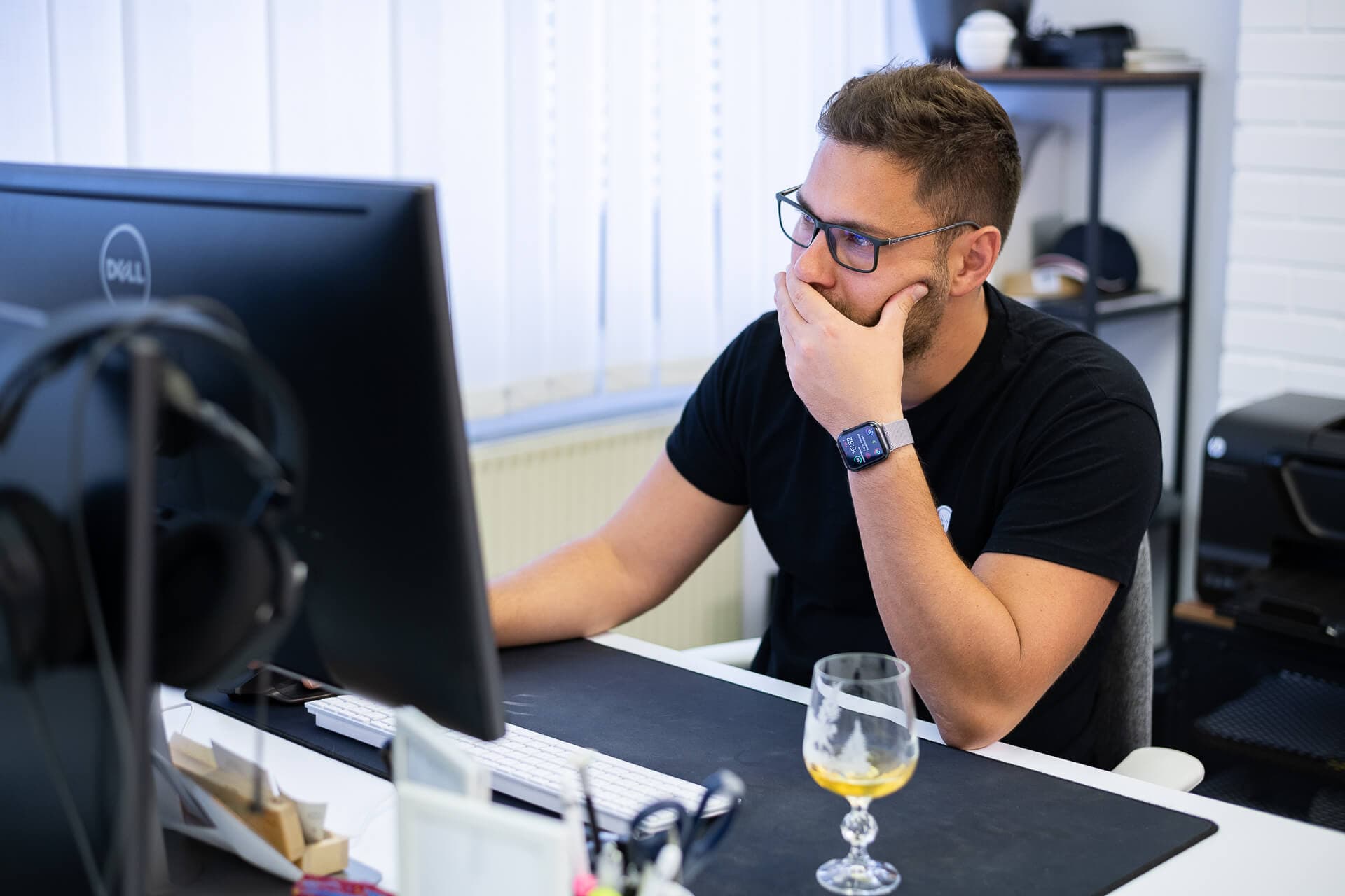 A man with glasses sits at a desk, looking thoughtfully at a computer screen while resting his chin on his hand. A glass containing a yellow beverage is placed on the desk, alongside a few items like papers and a headset. The office has white walls and natural light coming through the blinds.