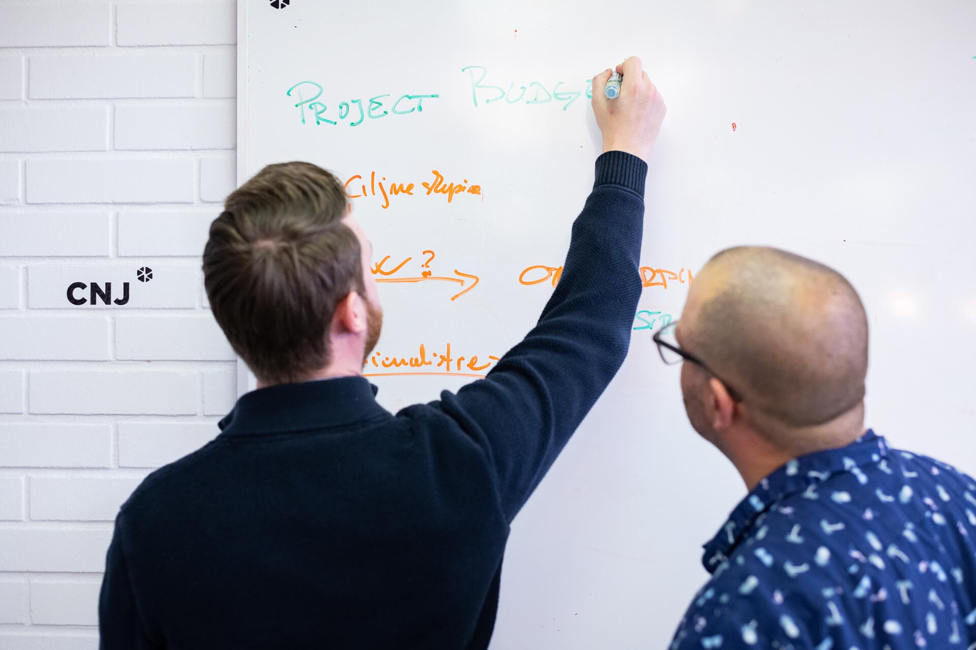 Two people are collaborating in front of a whiteboard, one writing with a marker while the other observes. The whiteboard features notes, including "PROJECT BUDGET" and arrows pointing to other items. The individuals appear focused on planning or discussing a project.