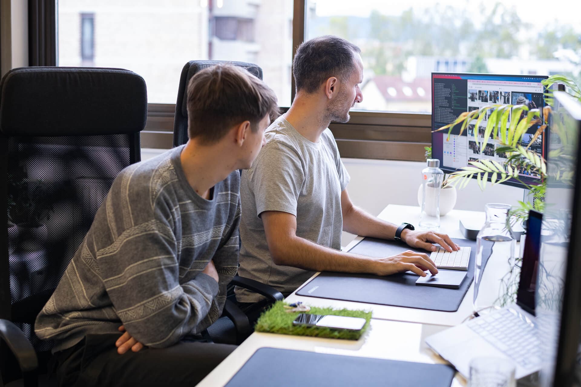 Two men are seated at a modern office desk, one actively working on a computer while the other observes. The workspace includes multiple monitors, a water bottle, and a small plant on the desk, with natural light streaming in through a window.