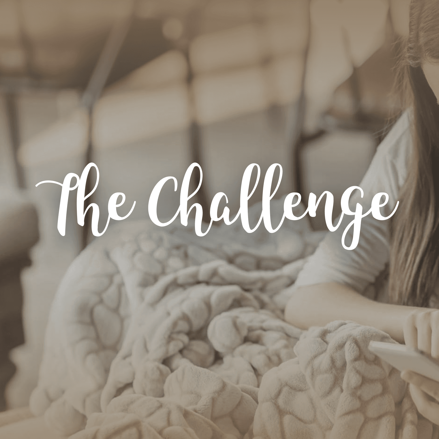 Young woman wrapped in a soft, light gray blanket sitting on a couch, holding a tablet. Overlay text in white cursive font reads "The Challenge." Background shows blurred chairs and warm lighting, evoking a cozy and relaxed atmosphere.