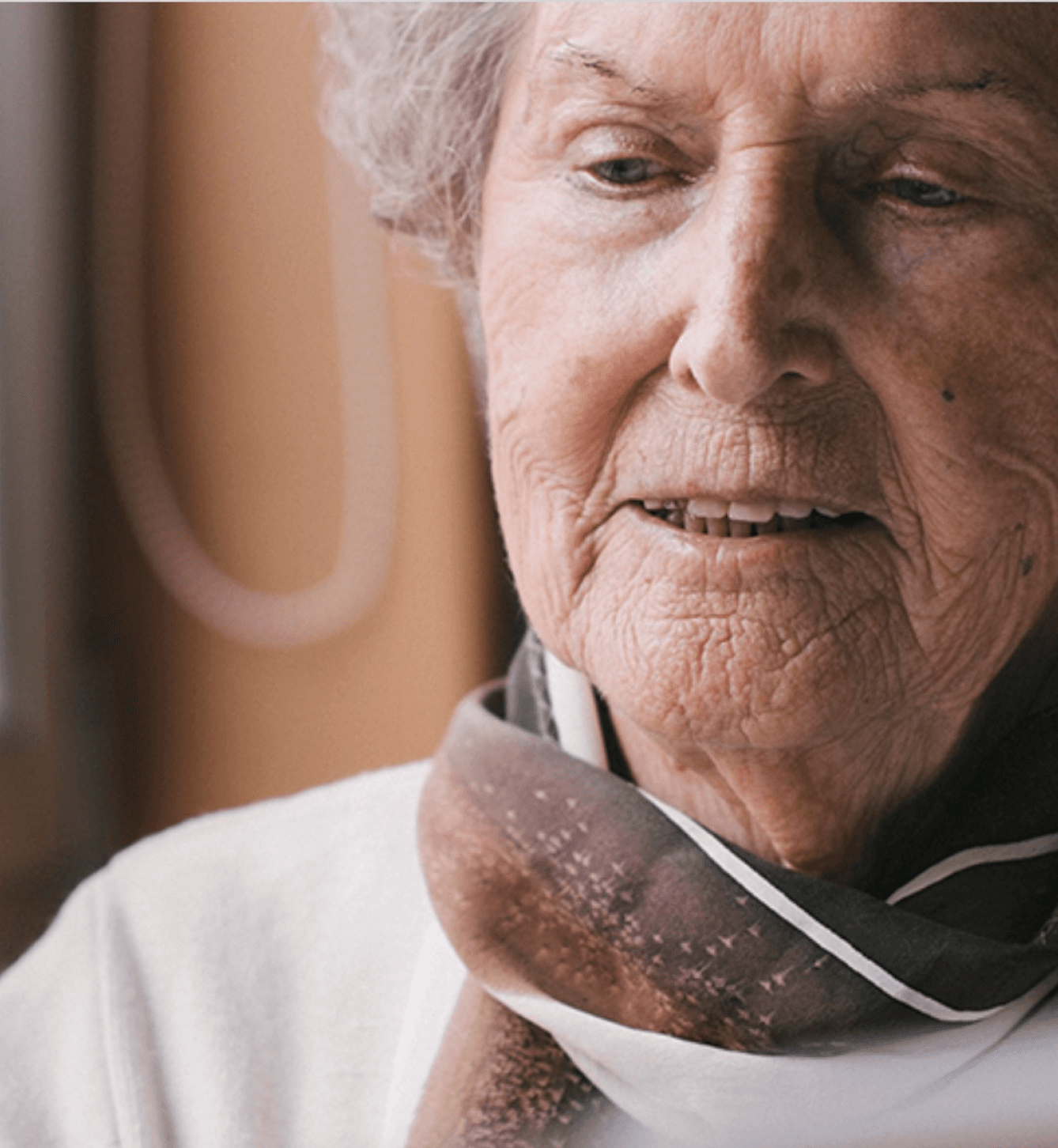 Elderly woman with short gray hair and light skin, wearing a patterned scarf and white top, looking downward with a gentle expression in natural light.